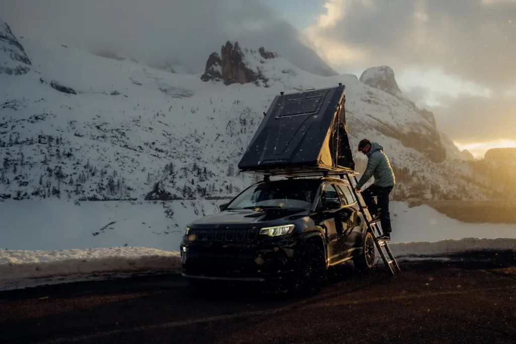 Mann klettert in ein geöffnetes Dachzelt auf einem 4x4 SUV im winterlichen Gebirge.