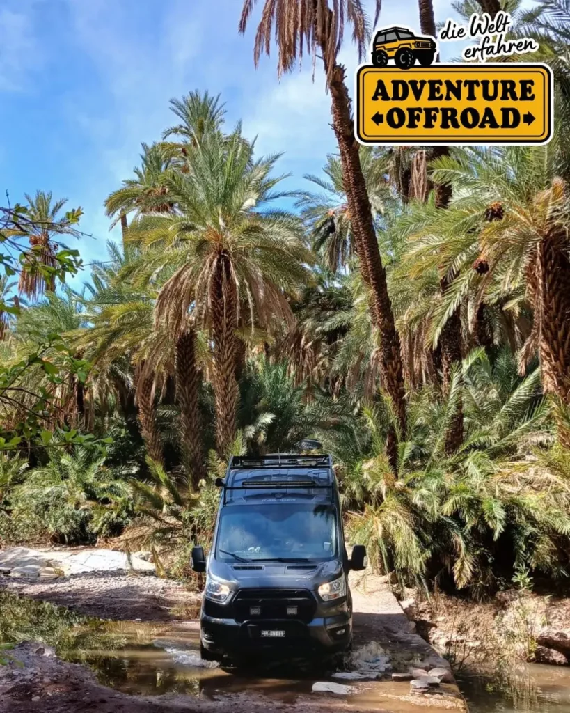 Schwarzer 4x4 Campervan mit Dachgepäckträger durchquert ein steiniges Flussbett in einer Palmenoase.