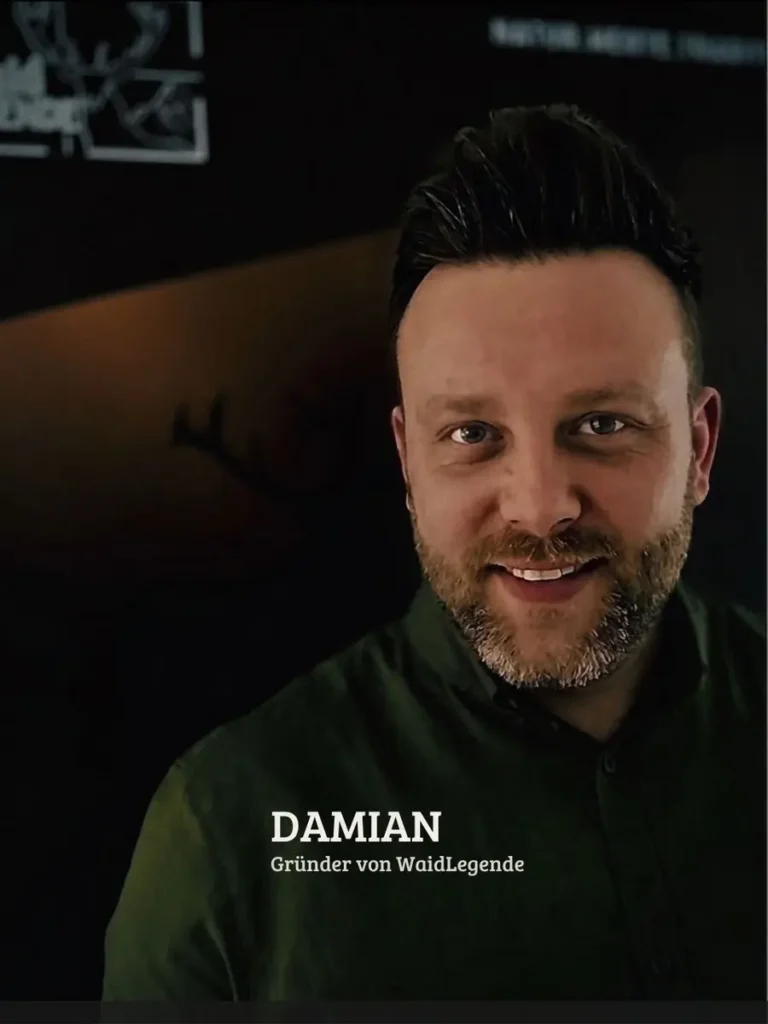 Ein lächelnder Mann mit Bart und dunklem Haar trägt ein olivgrünes Hemd vor dunklem Hintergrund.
