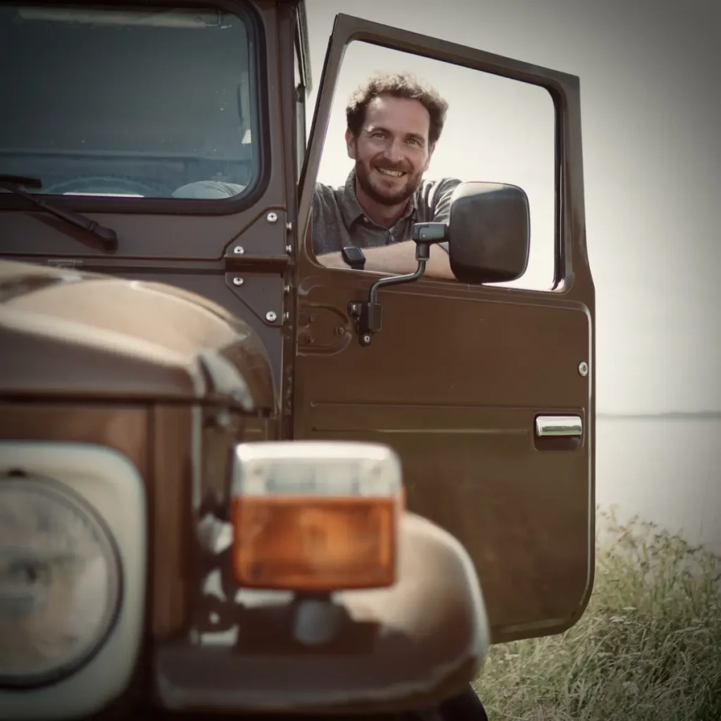Ein bärtiger Mann lächelt aus einem braunen 4x4 Geländewagen mit See im Hintergrund.