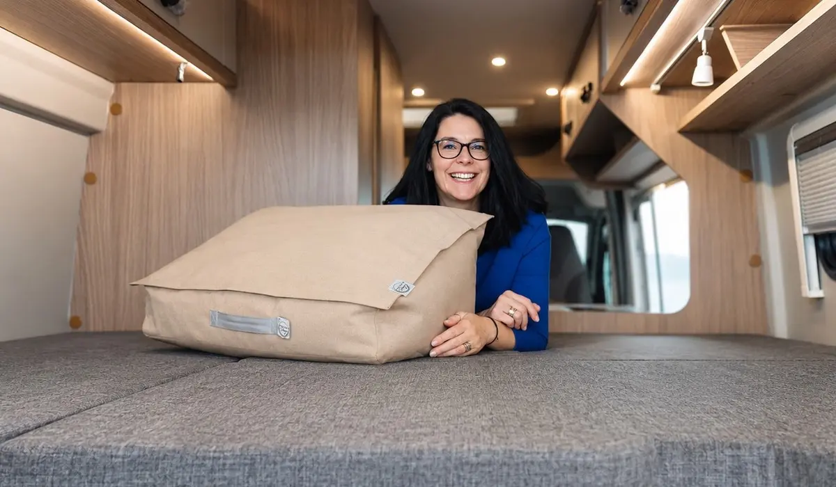 Eine lächelnde Frau liegt im Campervan-Bett und zeigt eine große, beige Stautasche.
