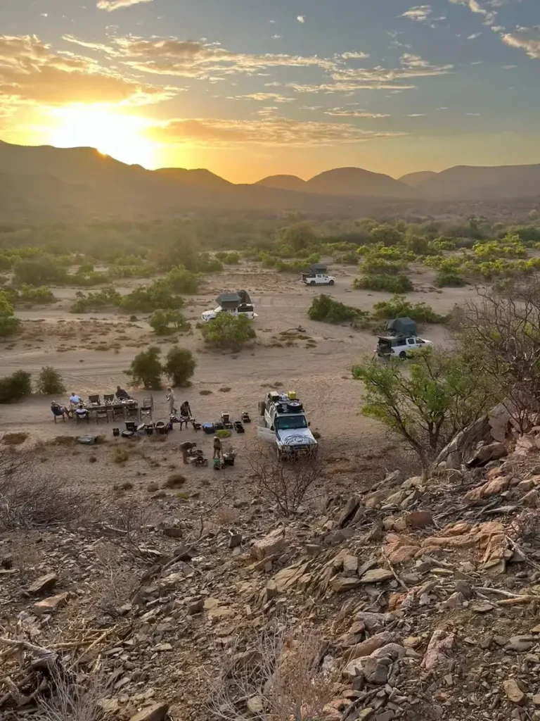 Mehrere 4x4-Expeditionsfahrzeuge und eine Gruppe Camper beim Sonnenuntergang in einer Wüstenlandschaft.