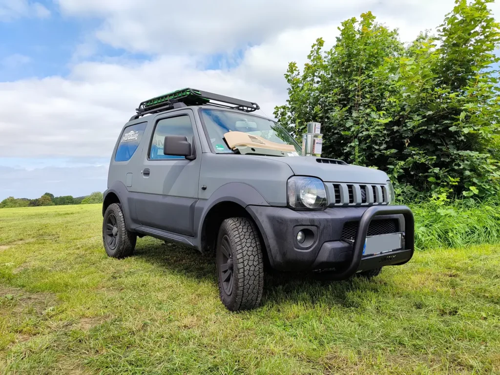 Ein grauer Suzuki Jimny 4x4 Offroad-Fahrzeug mit Dachgepäckträger und Frontschutzbügel auf einer Wiese.