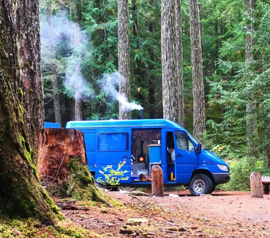 Blauer 4x4 Campervan mit offener Schiebetür und rauchendem Ofen steht im Wald.