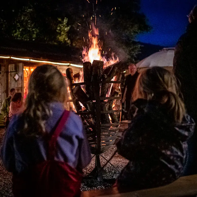 Zwei Personen sitzen abends vor einem großen Lagerfeuer in einer Feuerschale.