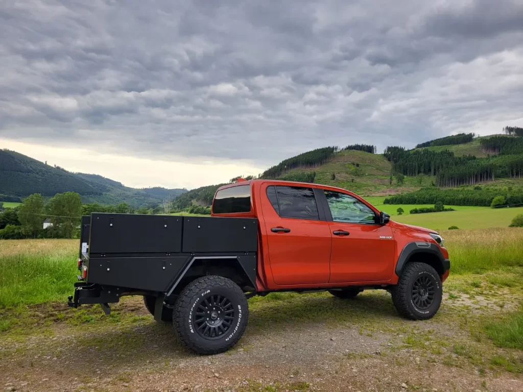 Ein orangefarbener Toyota Hilux Pick-up mit schwarzem Pritschenaufbau und Offroad-Bereifung steht in bergiger Landschaft.