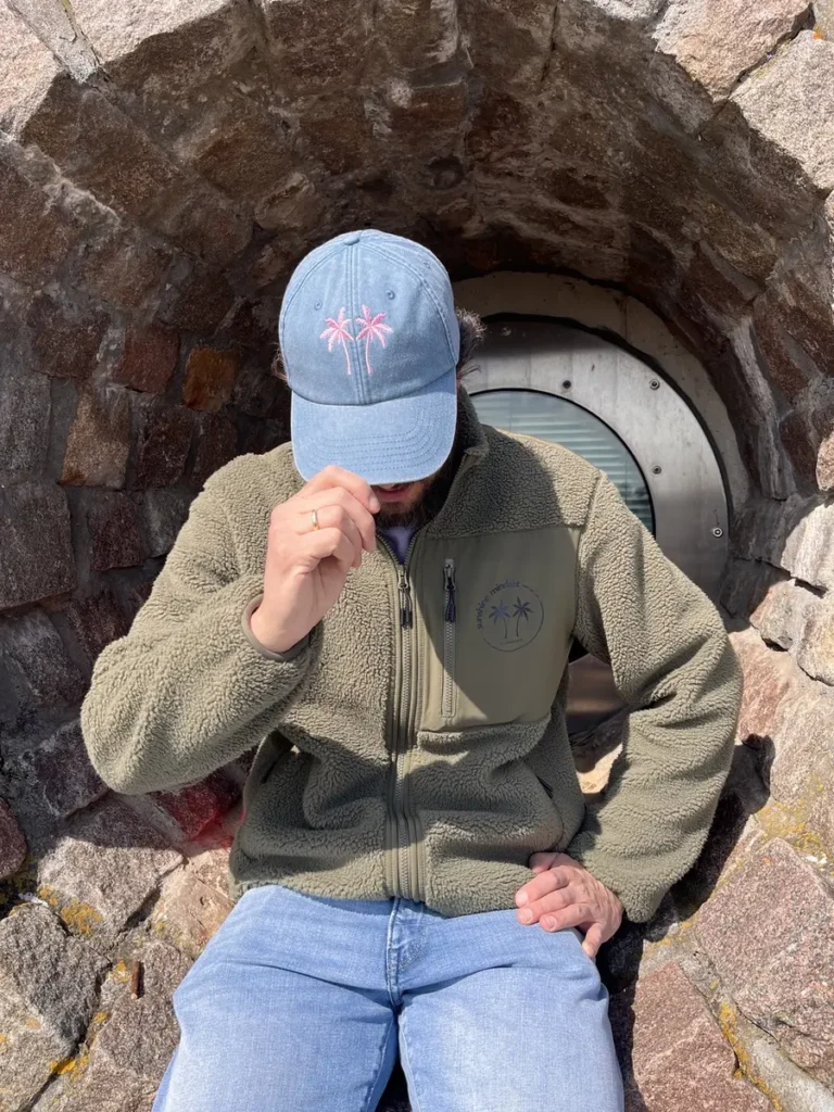 Ein Mann mit blauer Cap und Palmenmotiv trägt eine olivgrüne Fleecejacke vor einer Steinwand.
