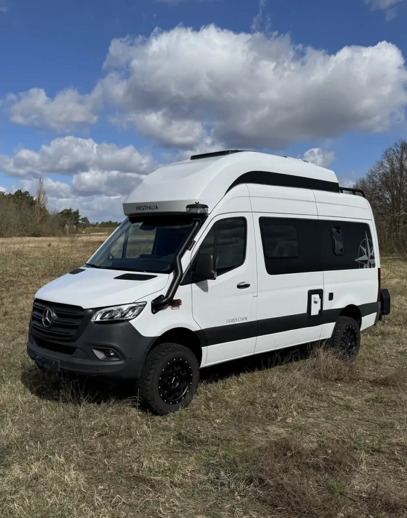 Weißer Westfalia James Cook 4x4 Campervan mit Geländeausstattung steht auf einer Wiese.