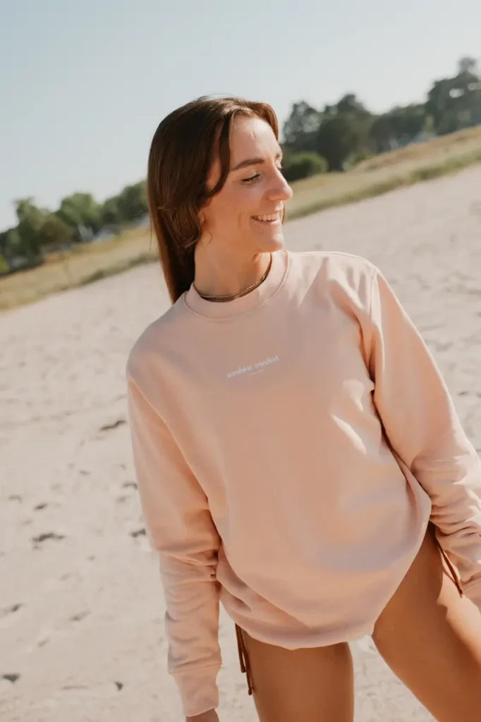 Eine junge Frau im hellrosa Pullover lächelt am sonnigen Strand.
