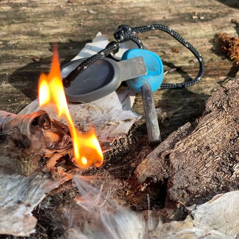 Ein Feuerstahl mit Schlagplatte liegt neben brennender Birkenrinde und Zunder.