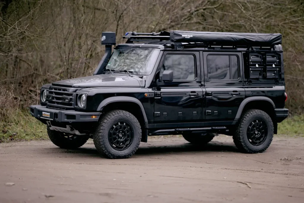 Schwarzer Ineos Grenadier Geländewagen mit Schnorchel, Dachmarkise, Offroad-Reifen und Heckstauraum auf unbefestigtem Weg.