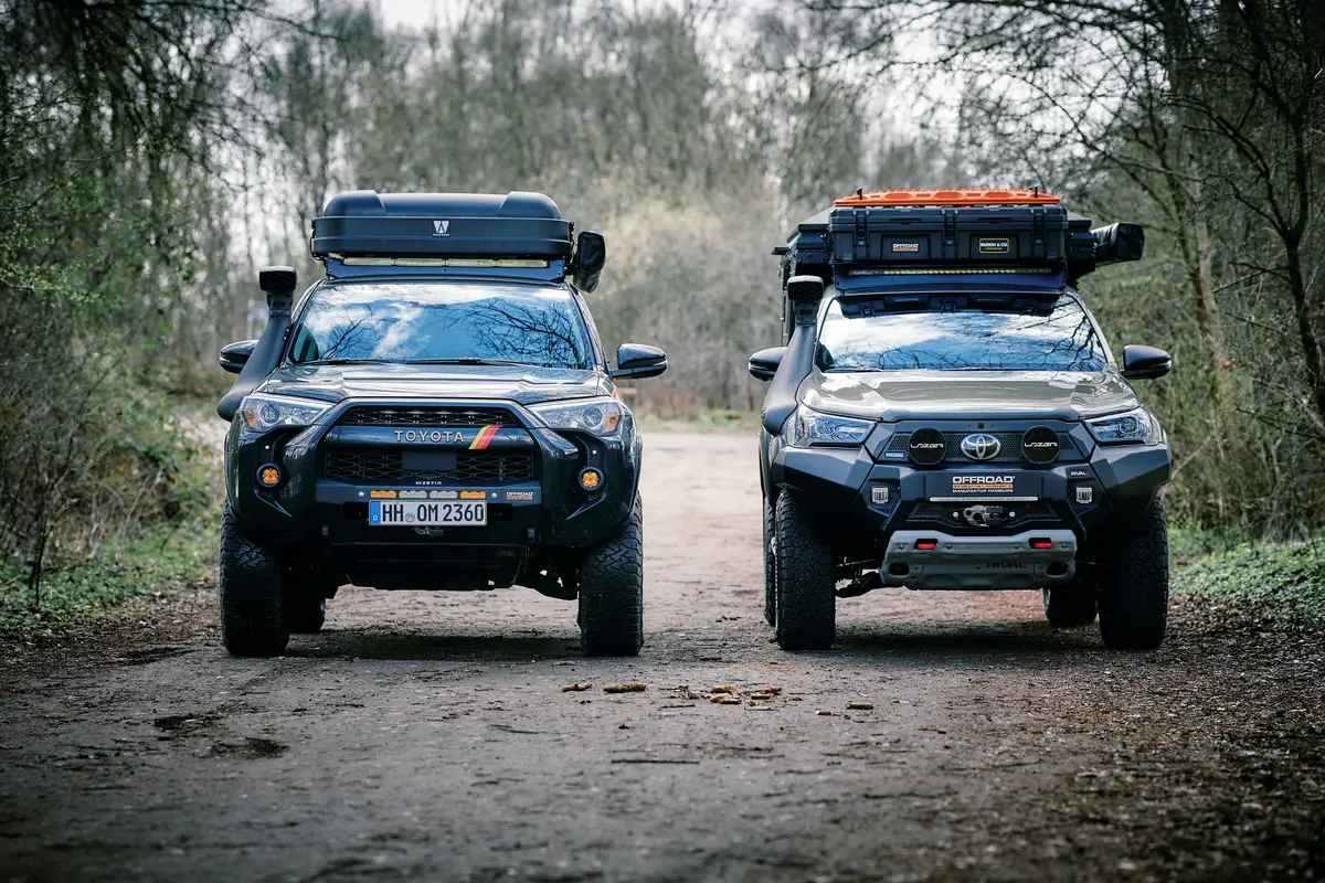 Zwei stark modifizierte Toyota 4x4 Offroad-Fahrzeuge mit Dachgepäck und Snorkel auf Waldweg.